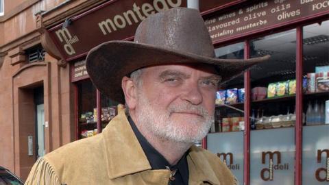 Angus MacInnes looking straight at the camera. He is wearing a brown cowboy hat and has a short, white beard. He is wearing a light brown suede jacket with frilled sides and is standing in front of a shop on the set of River City.