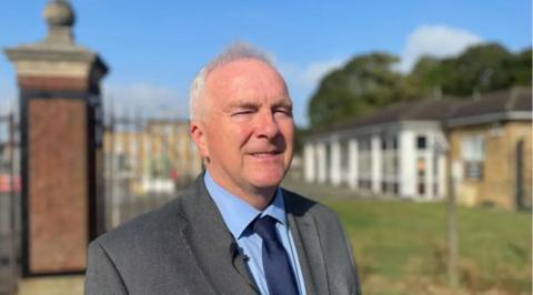 Trevor Young standing in front of a building wearing a grey suit, a blue shirt and a blue tie