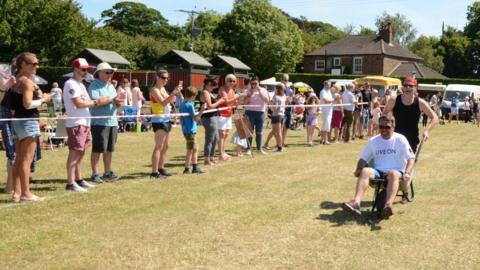 A woman pushing a man in a wheelbarrow along a grass field, with spectators applauding as they stand behind a white tape. Stalls and houses can be seen in the background.