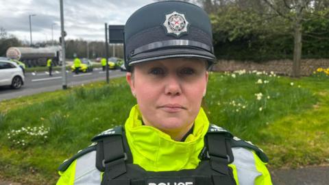 A police officer is wearing a high viz jacket. Her body protector says police. Her hat has the PSNI emblem.