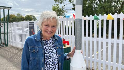 A woman standing on a platform at a railway station. She has short white hair and is wearing a patterned top and a blue denim jacket. There are white railings behind her. 