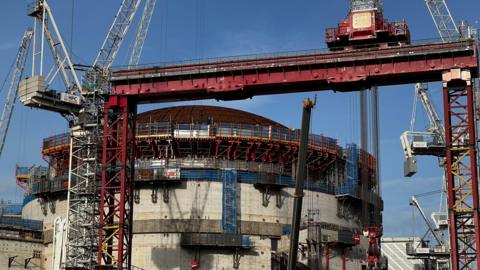 A wide cylinder building in the background, with a domed roof surrounding by cranes and giant structures. In the foreground is a red cargo-style lift.