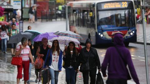 Several people walk along a pavement in the rain wearing hooded jackets and carrying umbrellas. A single decker bus is on the road in the background.