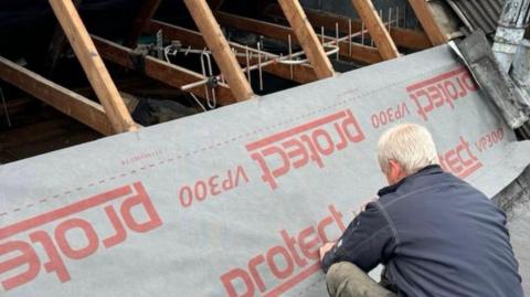 A man with white hair is kneeling whilst working on a section of a roof, where wooden supporting beams can be seen.