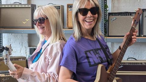Two members of the band Nanaz pose with guitars, standing back-to-back. They're wearing sunglasses and smiling to camera. 
