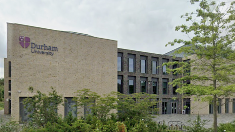 Durham University's teaching and learning centre is a multi-storey building made of beige bricks. It has a section of tall windows at its centre. There is a courtyard in front of the building, as well as some shrubs and young trees. The university's grey and purple logo is placed near the top of the building.