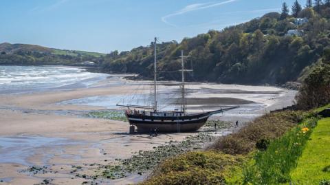 A traditional tall ship sits upright on a sandy beach with seaweed-covered rocks around it. To the right and behind is a hillside covered in trees and fields around the jagged coastline, with the tide out. It is a sunny day.