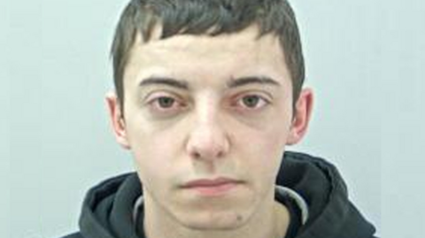 A mug shot of Dylan Green. He has short dark hair, is wearing a dark jacket. He is staring into the camera lens.