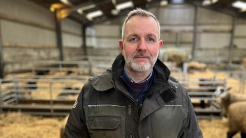 Michael Goldie looking at the camera, wearing a khaki jacket. He is standing in a barn with his surviving lambs and ewes in pens behind him.