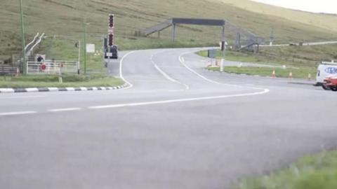 A road snaking up a hill with a metal footbridge over the carriageway