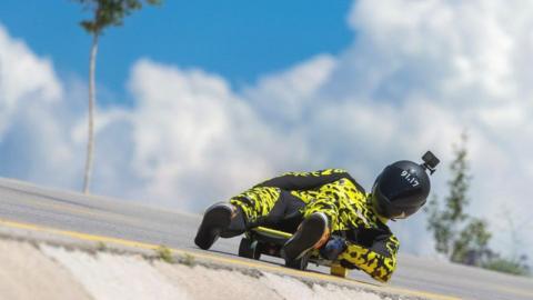 Phil Spencer in race suit and crash helmet downhill skateboarding in Turkey. The sky is blue behind him, with the race suit bright yellow and black. 