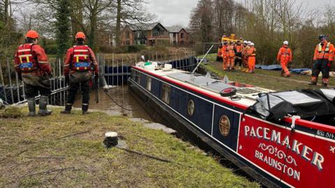 dark blue and red canal boat in a small section of canal blocked off with metal dams. it is in water and there are dozens of people standing on banks either side of it in orange outfits