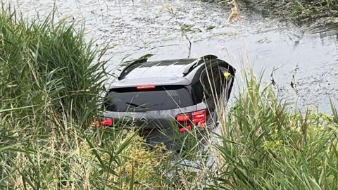 A silver and black car has entered a river which has several bits of grass floating on it. The river bank is on the left side of the image.