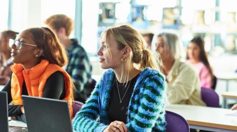 A room of students sitting at desks, most are blurred but two women at the front, one has red/orange long hair and is wearing glasses and a black long sleeved top with an orange gilet, the other woman has blonde long hair tied in a ponytail, she is wearing a blue and green striped jacket and a black top with a silver necklace. She is also wearing hooped gold earrings. 