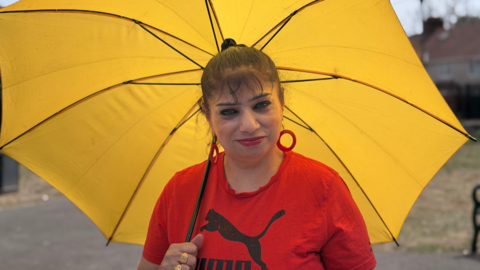 A woman wearing a red Puma top and red circular earrings underneath a yellow umbrella. She is smiling into the camera.