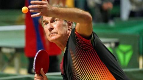 John Poysden staring at an orange table tennis ball he has just thrown in the air. He is holding a red paddle and a black and red top.