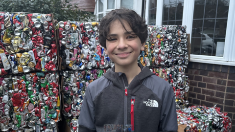 A boy with dark hair and wearing a grey top is looking at the camera. Many cans are stacked behind him in separate blocks outside a property, with windows on the top right of the photo.