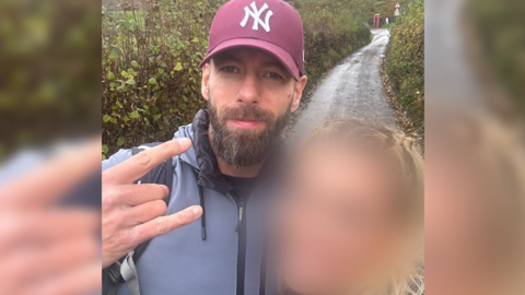 John Hall is holding his fingers up to the camera with a slight smile on his face. He is wearing a purple baseball cap and grey water proof jacket. His victim is standing next to him with her face blurred out. They are standing on a narrow road with bushes either side. 