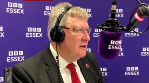 Roger Hirst, wearing headphones talking into a BBC Essex microphone, with a BBC Essex backdrop behind him