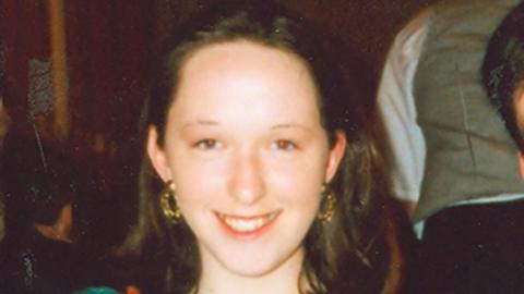 A woman, pictured from the neck up, smiling at the camera. She has long brown hair and is wearing earrings. 