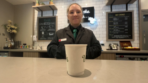 Sorcha Calvert stands behind a coffee bar at the Leonardo Royal Hotel Oxford - a coffee cup can be seen on the bar in the foreground. She is dressed smartly in a waistcoat, shirt and green tie and is smiling at the camera.
