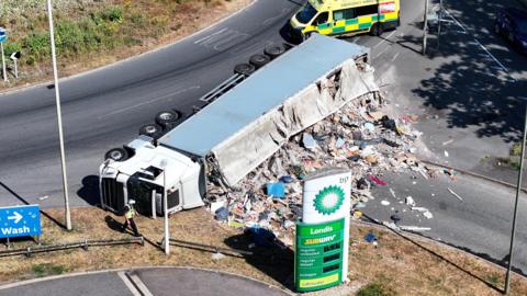 A white lorry is on its side next to a roundabout. What appears to be mounds of rubbish is coming out of the lorry. In the foreground is a white and green BP advertising pillar. Behind the lorry is a yellow and green ambulance parked in the road