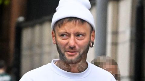 A man with a short beard, neck tattoos, two piercings below his bottom lip and a large circle earring in his ear wears a white beanie. He also wears a white sweatshirt as he walks through a street.