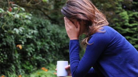 A woman is sitting outdoors in a garden, holding a mug and covering her face with one hand, with greenery and flowers in the background.
