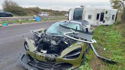 A green damaged McLaren 720S car is parked on the side of the road, a large section of the front of the car is destroyed and the windscreen is smashed. 

Behind it is a white motorhome vehicle laying on its side.

Debris can be seen across the road.