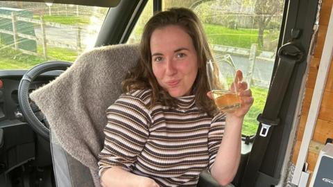 A woman with mid-length brown hair sat inside a van drinking a glass of juice, eating her breakfast 