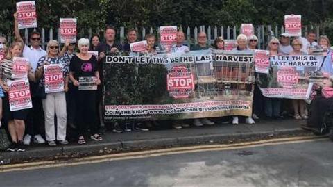 A group of people standing on a footpath holding signs and protesting against a proposed housing development. 