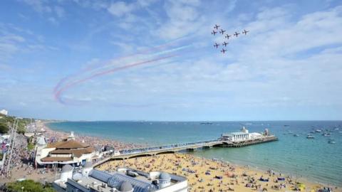 A view of Bournemouth beach and pier, with the Red Arrows flying in formation overhead. The beach and promenade area are packed with people.