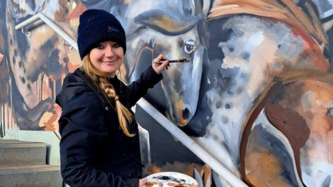 A woman stands in front of a wall with a large mural painted on it. She has a long blonde plait and she is wearing a black hat and a black hoodie. She is holding a paintbrush and a tray with paint on it. The mural has grey horses on it.