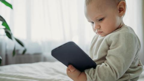 A baby in a grey jumper sits on a bed looking at a black phone.