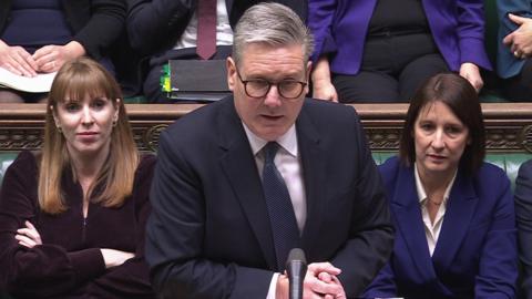 Keir Starmer in suit and tie standing in front of microphone, with hands crossed, with other MPs behind him