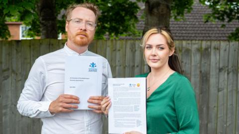 A man with ginger hair and a beard in a white shirt and glasses is holding a piece of paper that says "justice" in blue ink. A woman with a blonde fringe and brown hair is wearing a silky green shirt and golden heart necklace. She is also holding a piece of paper with a lot of printed text on it. Both are stood outside in front of a wooden gate.