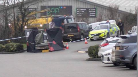 The scene where the car was found. A silver car has been surrounded by black boards and tarpaulin but the back of it is still visible. There is a parked police car and two policemen as well as police scene tape stretched around the car via traffic cones. A metal warehouse-type building is in the background with a sign reading GAP.