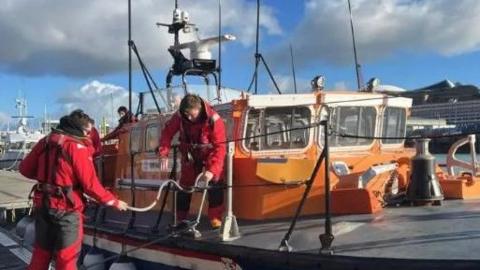 Two crew members in red outfits next to an orange lifeboat