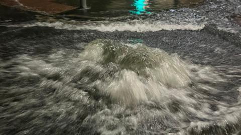 Water surges out of a manhole cover on a road. 