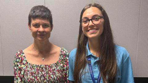 Two NHS Somerset nursing apprentices are standing next to each other in front of a white wall. The left woman is wearing a colourful dress and has short hair. The woman on the right is wearing a blue shirt and has long, dark hair.