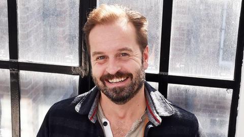 Head and shoulders image of a smiling Alfie Boe standing in front of a window with small black framed panes. He has short dark hair and a beard and is wearing beige button up top under a navy shirt jacket