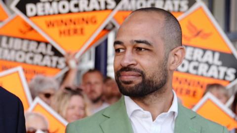 Josh Babarinde MP campaigning in Eastbourne at the general election in July 2024. Behind him are a series of Liberal Democrat campaign signs.