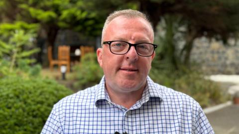 Group chair Garin Dart. He is wearing a white and blue check shirt. He has short balding, blonde hair and black glasses. 