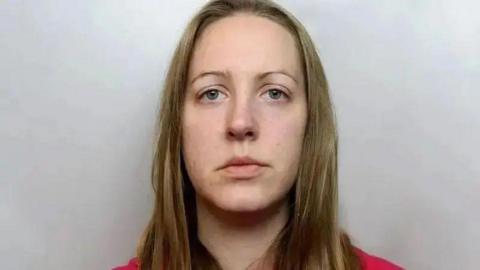 Custody photograph of Lucy Letby. She has long blonde hair and blue eyes, is wearing a red top and is looking straight into the camera lens