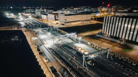 Aerial view of Cambridge South station at night-time. There are construction workers on the railway tracks.