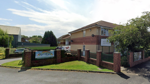 The Gables Care home is a two-storey brick building on a residential road. The main building is slightly hidden behind trees but a sign reads The Gables Care Home.