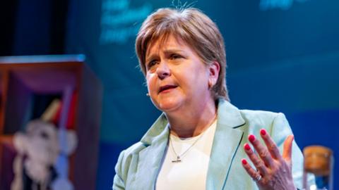 a woman with short brown hair is wearing a green jacket over a white t-shirt and has her left hand out in front of her