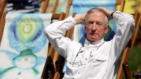 Author Raymond Briggs sat on a deck chair. He the deck chairs have drawings of The Snowman on them.