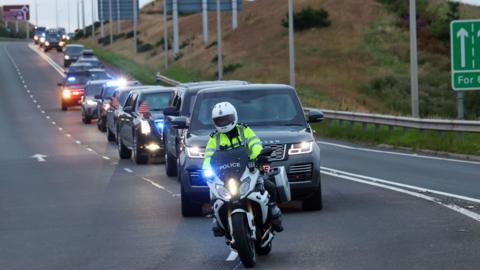 A large line of vehicles led by a police motorbike, drive along the road 