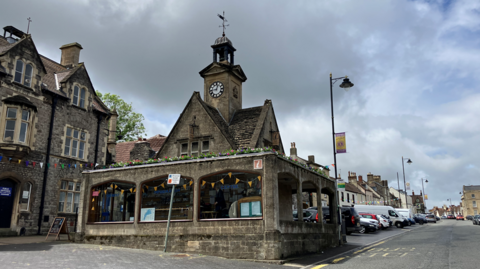 A stone clock tower, with a weather vane on top, stands at the edge of a high street. There are flower boxes on the upper level. Its lower section features a partially glazed arcade. Rows of parked cars can be seen in the distance and to the left is a three storey brick building.